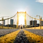 Suspension bridge with yellow paint, backlit by the sun, leading to a city skyline with high-rise buildings.