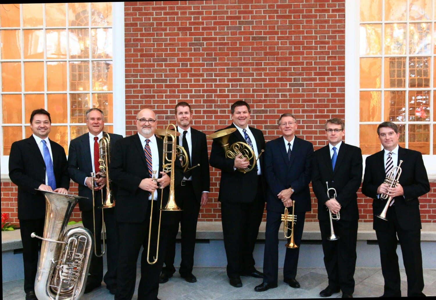 Eight men in suits holding various brass instruments stand in front of a brick wall with large windows.