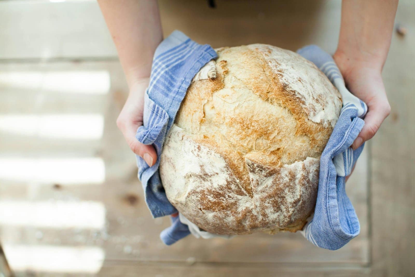 Hands holding a round loaf of rustic, crusty bread with a blue striped cloth.