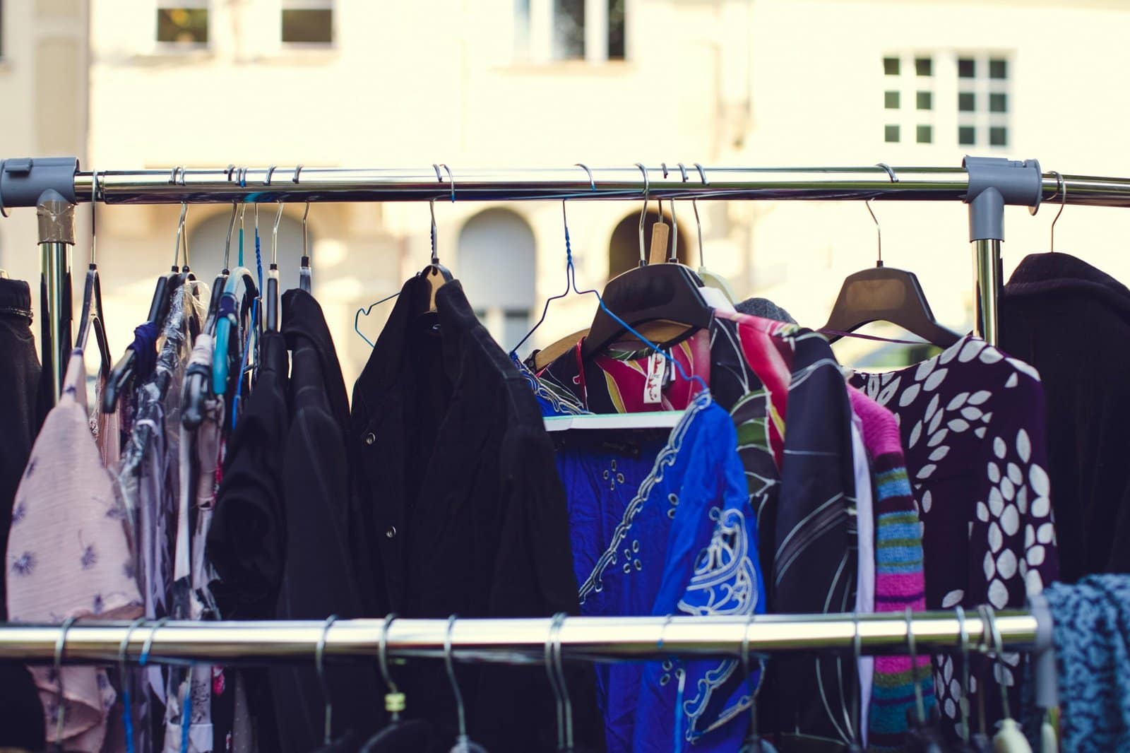 A clothing rack with various colorful garments on hangers, set outdoors with a blurred building in the background.