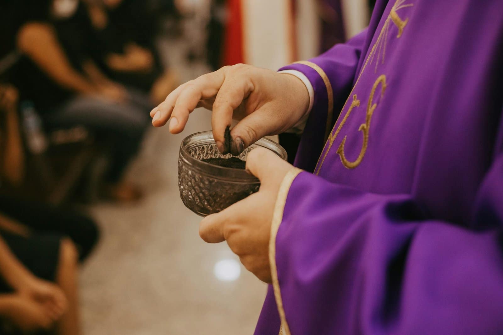 A person in a purple robe touches a small bowl of ashes with two fingers.